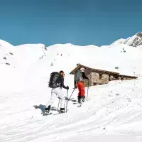 Deux skieurs en randonnée montant une pente enneigée vers un chalet en pierre en montagne aux Arcs