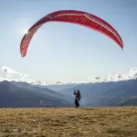 Un pilote de parapente prépare sa voile rouge au décollage sur une pente herbeuse face aux sommets enneigés des Arcs