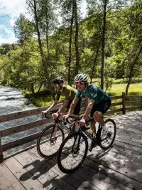 Deux cyclistes roulant côte à côte sur une passerelle en bois le long d’une rivière en forêt