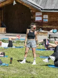 Une participante réalise un squat sauté énergique lors d'une séance de fitness de type HIIT en plein air aux Arcs, avec l'animateur en arrière-plan