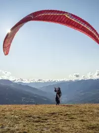 Un pilote de parapente prépare sa voile rouge au décollage sur une pente herbeuse face aux sommets enneigés des Arcs