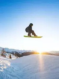 Un snowboardeur réalise un saut aérien impressionnant (grab) au snowpark des Arcs, avec le soleil couchant qui illumine les sommets de la Tarentaise