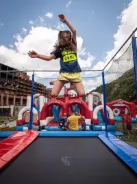 Une jeune fille en plein saut acrobatique sur un trampoline géant au Hero Camp d'Arc 2000, avec des structures gonflables en arrière-plan